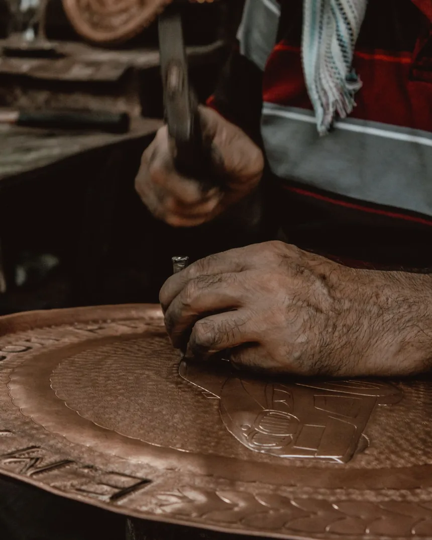 Detalle de una mano guiando el cincel y otra golpeando con el martillo sobre una chapa de cobre en relieve